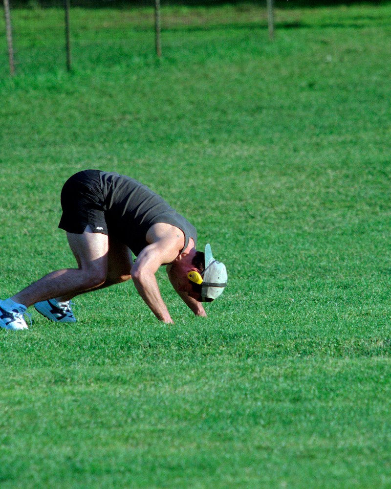 A man working out on all fours in a green field with a hat and headphones on