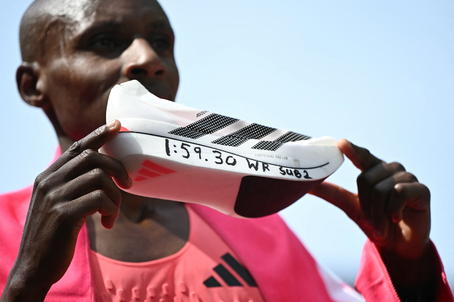 A man holding up a white running shoe with "1:59:30 WR SUB 2" written on the sole in black marker
