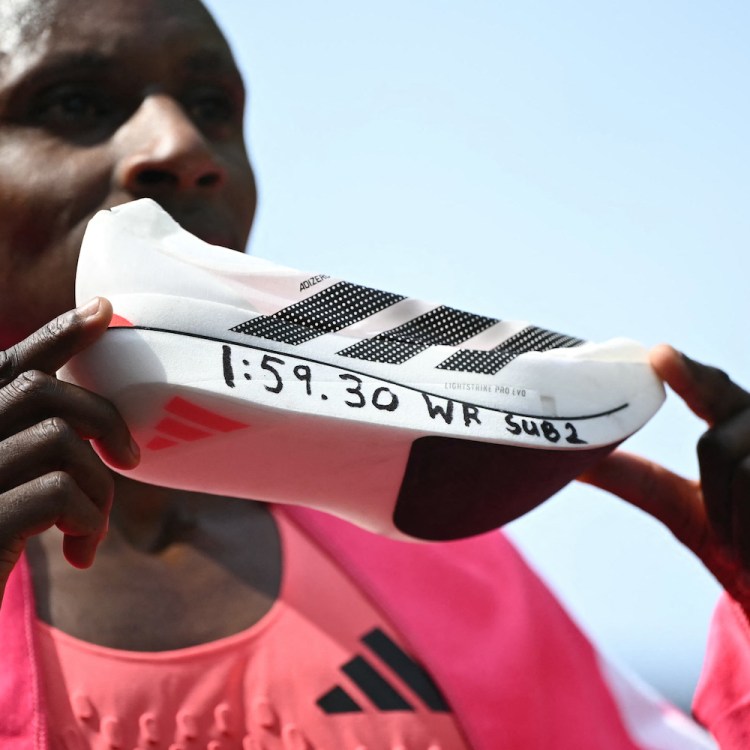 A man holding up a white running shoe with "1:59:30 WR SUB 2" written on the sole in black marker