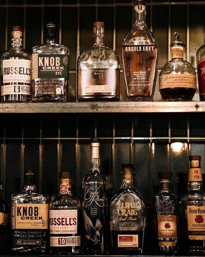 two rows of bourbon bottles sitting on a weathered metal backbar