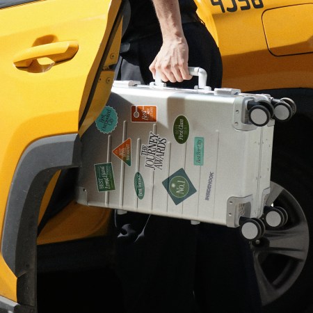 Man getting into a yellow taxi cab holding an aluminum suitcase covered in stickers