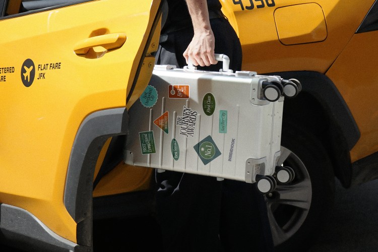 Man getting into a yellow taxi cab holding an aluminum suitcase covered in stickers