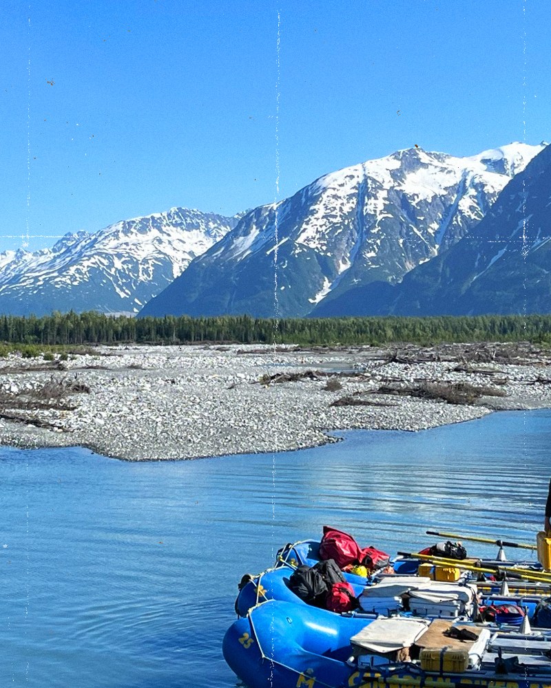 A person stands between two loaded blue rafts numbered 24 and 26 at a sunny river launch site, with a Momentum Alaska raft visible nearby. A braided glacial river winds through a rocky gravel bar, backed by a dense spruce forest and dramatic snow-capped peaks under a clear blue sky.