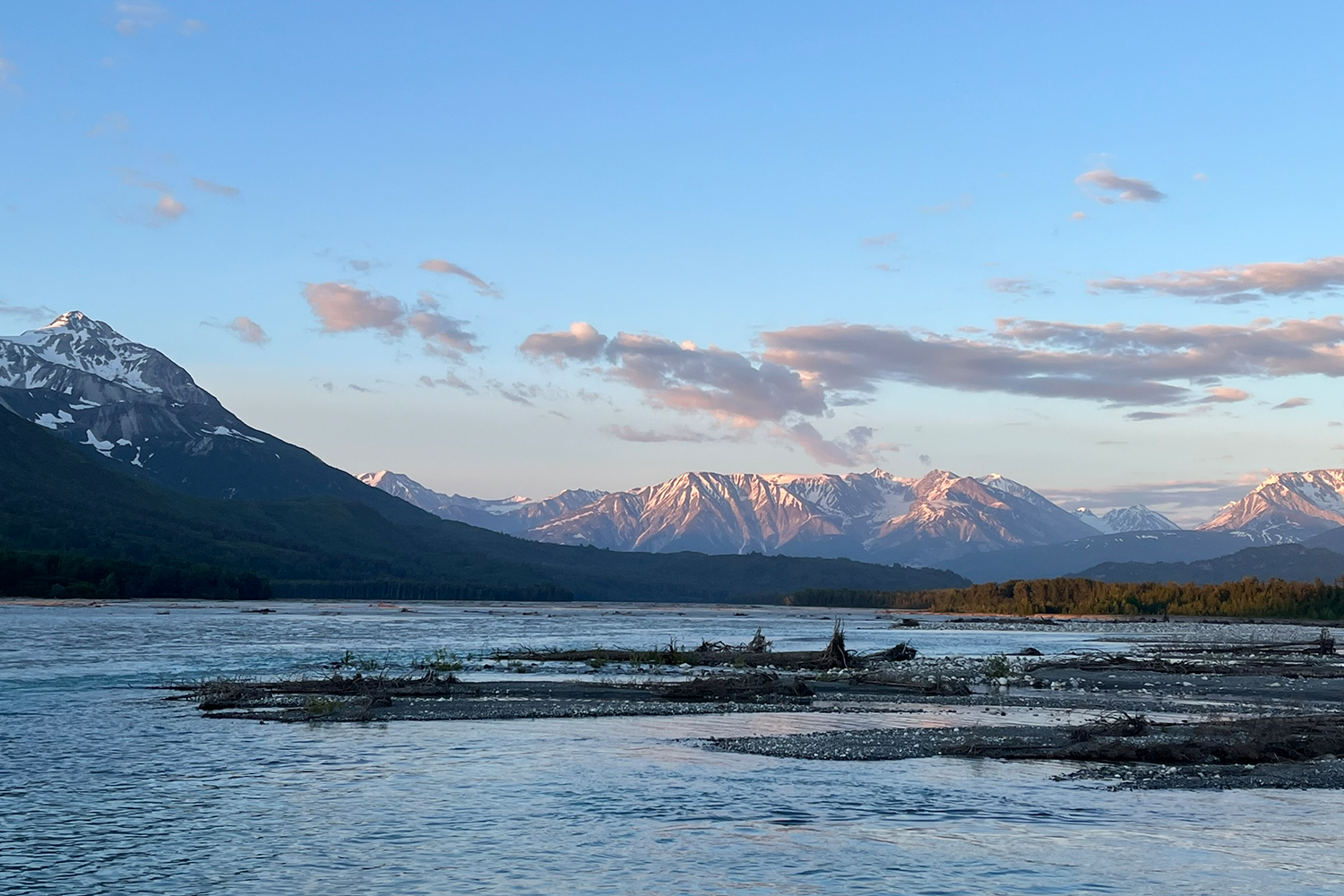 A wide, braided river flows past a driftwood-covered gravel bar at dusk, with golden-lit forest islands in the middle distance and a panoramic range of snow-capped mountains glowing pink and amber under a soft blue sky scattered with wispy clouds.