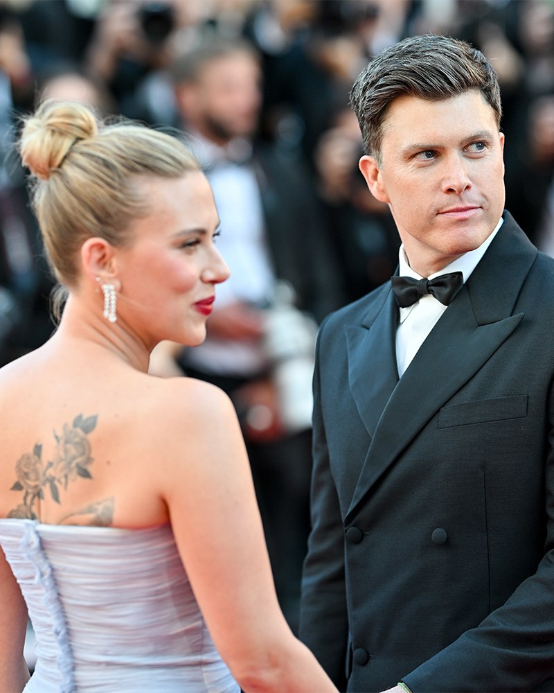 A man in a black tuxedo on a red carpet with a woman in a purple dress with her back turned