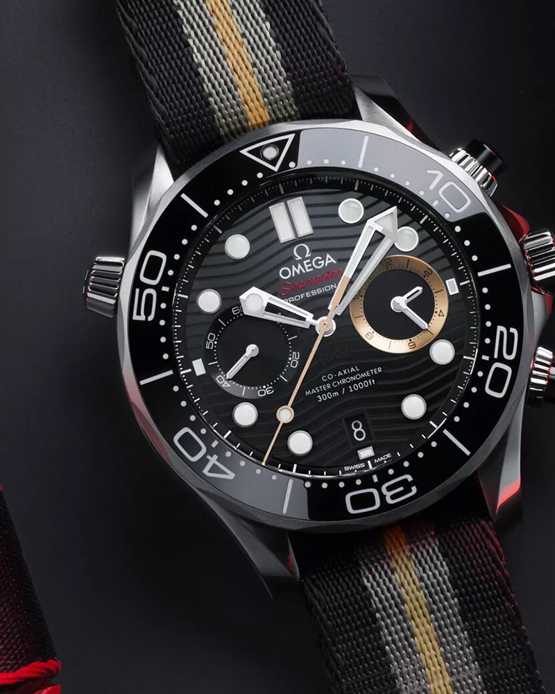 A watch with white hands, a black dial and bezel, and one gold subdial sits on a black table next to folded watch straps