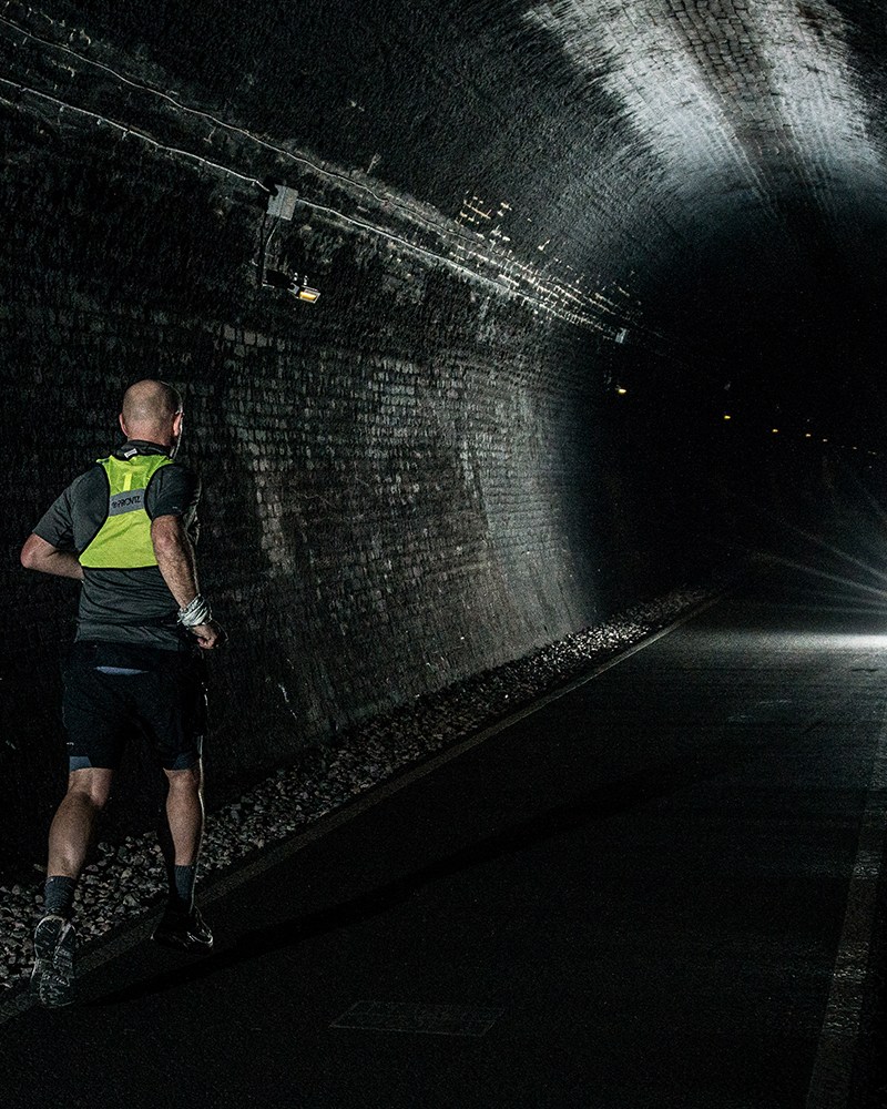 A man running in a high-vis vest through a dark tunnel with a single light shining in the distance