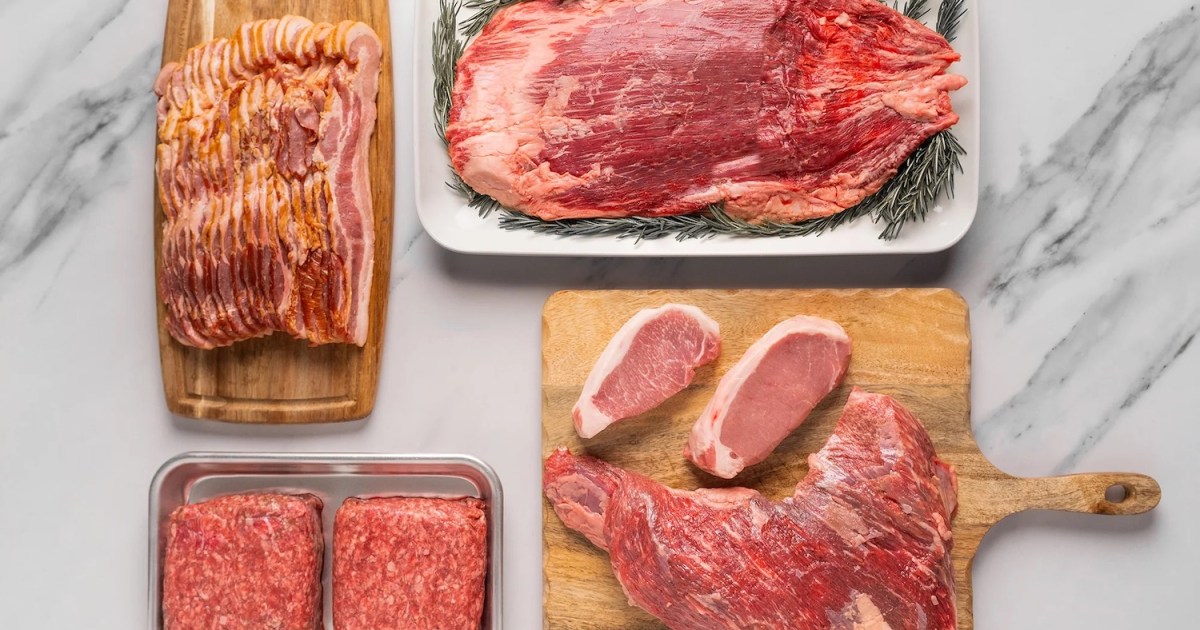 ground beef, pork chops, flank steak and bacon on trays and wooden cutting boards on a white marble surface