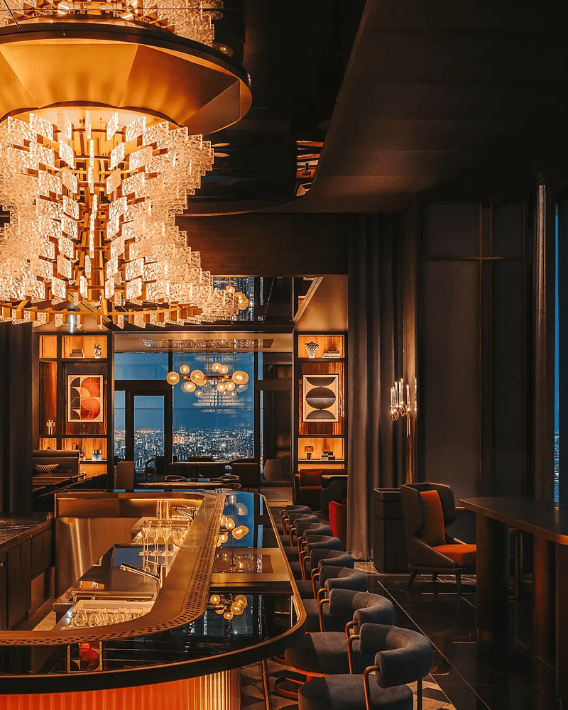 The warmly lit bar at Driftwood Bar & Restaurant in the Fairmont Tokyo, with a cascading crystal chandelier overhead, a curved black marble bar stocked with spirits and floor-to-ceiling windows overlooking the glittering Tokyo skyline at dusk.