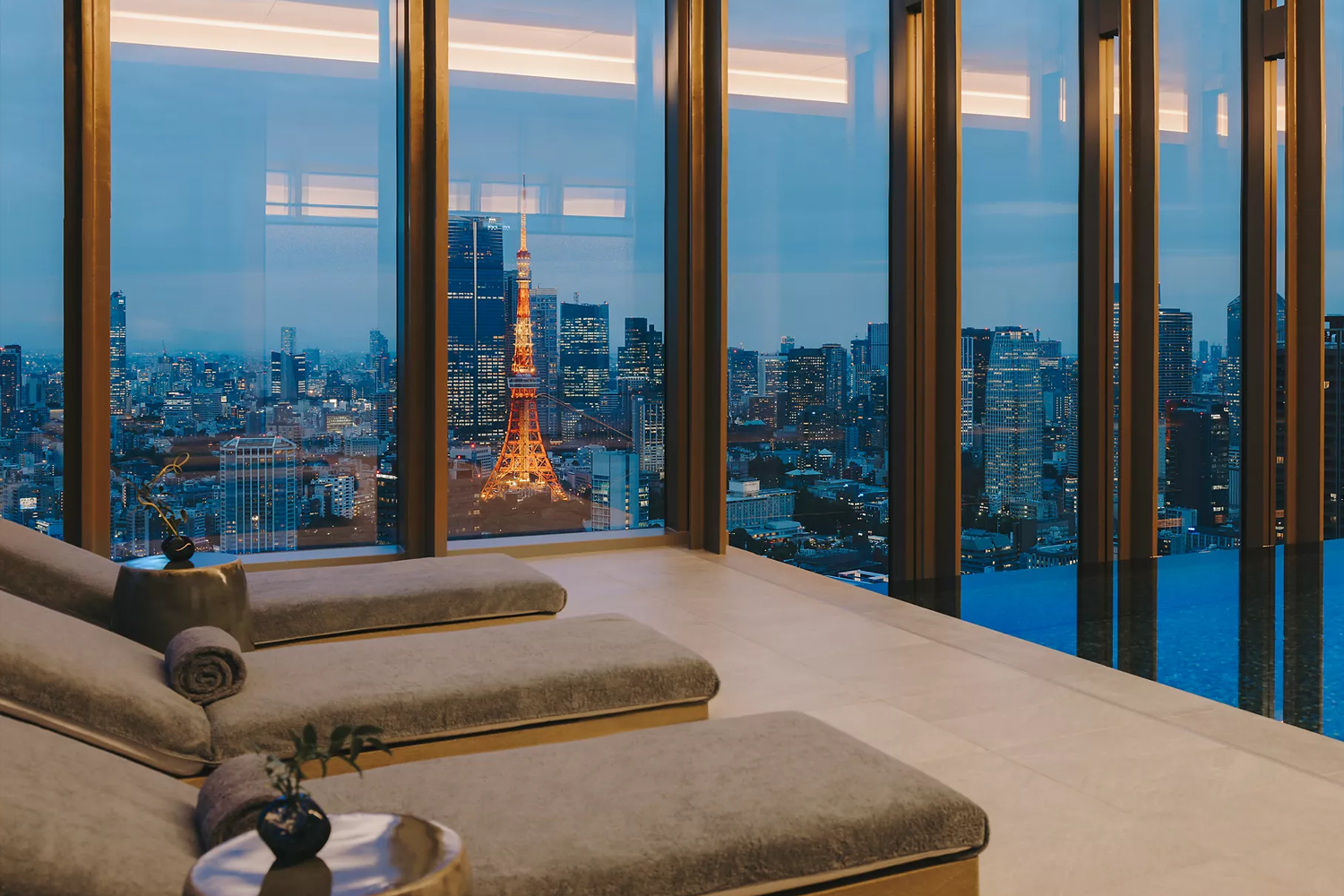 Interior of a hotel spa relaxation room at dusk, with two padded lounge beds in the foreground and floor-to-ceiling bronze-framed windows overlooking the illuminated Tokyo Tower and city skyline at blue hour.