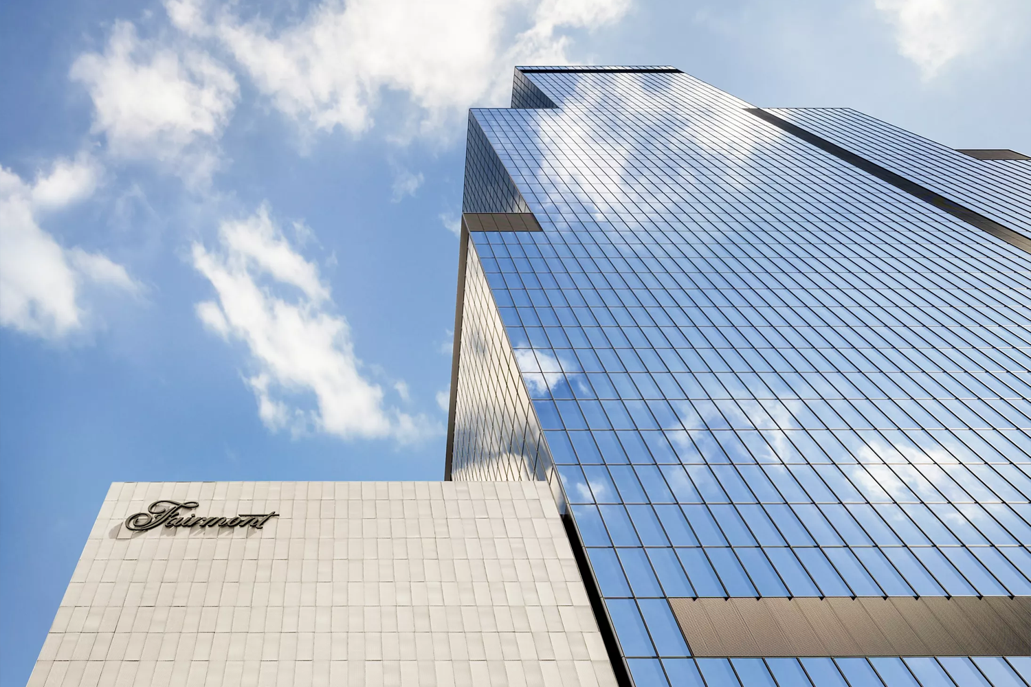 Low-angle exterior shot of the Fairmont Tokyo, showing a soaring glass-curtain-wall tower reflecting clouds and blue sky, alongside a lower cream-stone podium bearing the Fairmont script logo in black.
