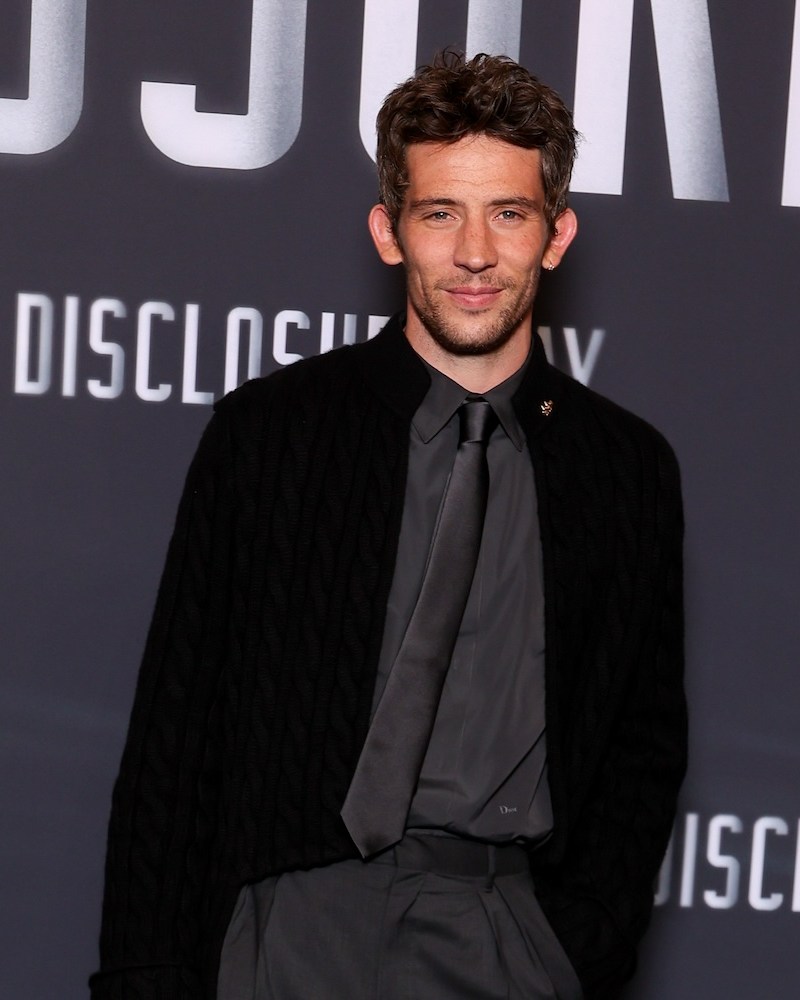 A man in a black cardigan, dress shirt and tie standing in front of a "Disclosure Day" backdrop