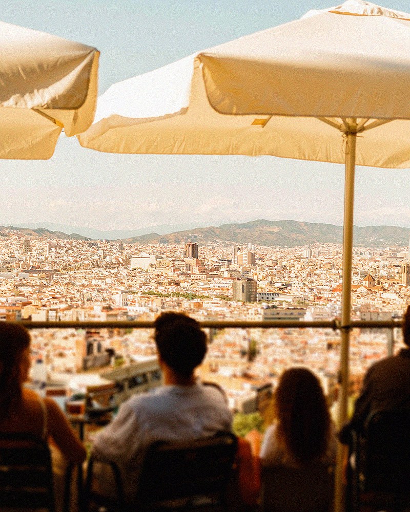 Travelers seated at an outdoor terrace café beneath white umbrellas, looking out over a sweeping panoramic view of Barcelona's skyline and rooftops, with mountains in the distance.