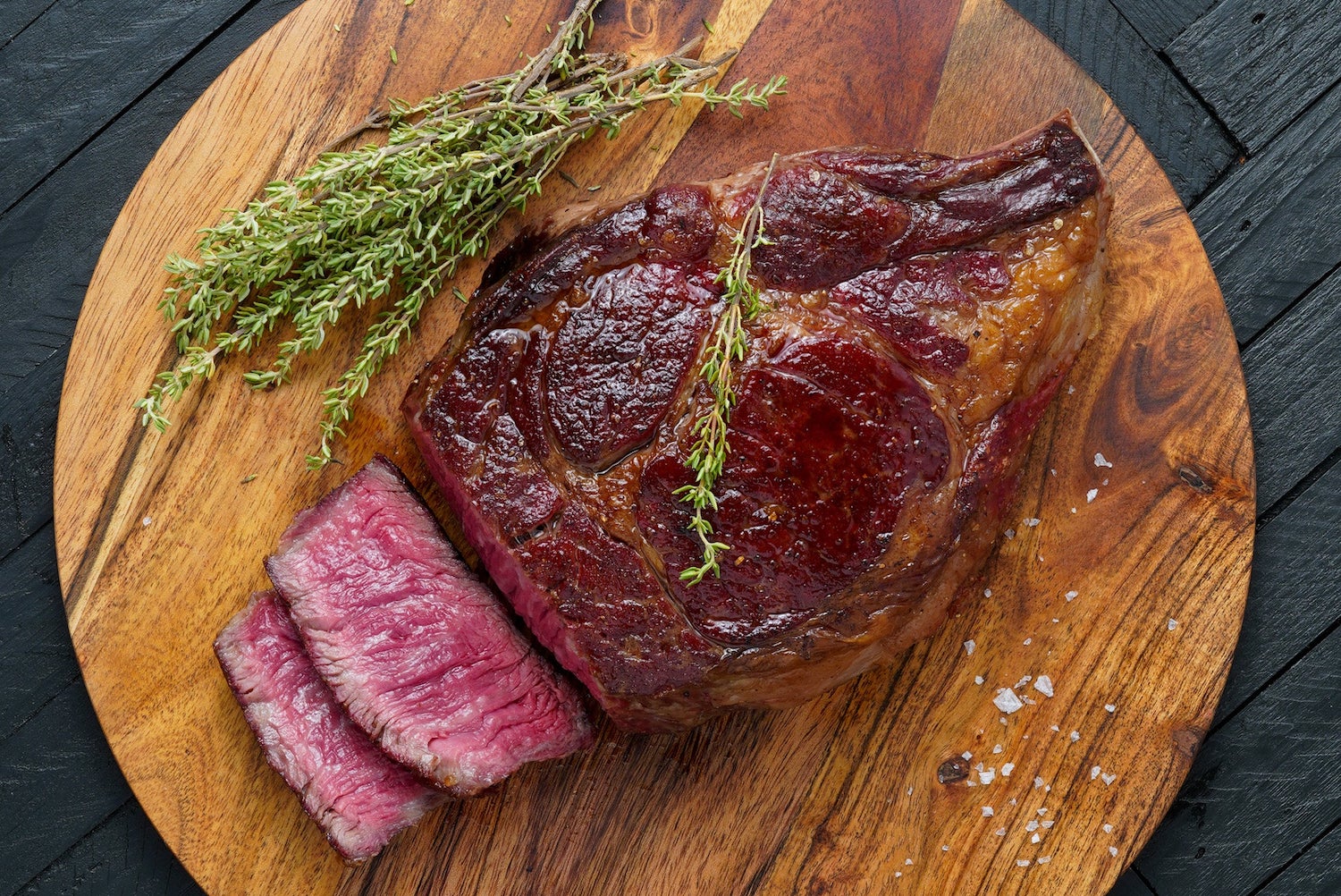 a ribeye with two slices next to it and thyme on a wooden cutting board