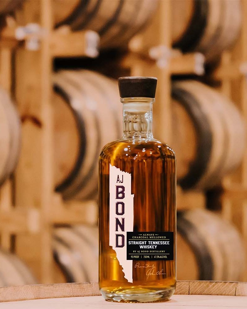 A bottle of AJ Bond whiskey in front of rows of barrels in a rickhouse