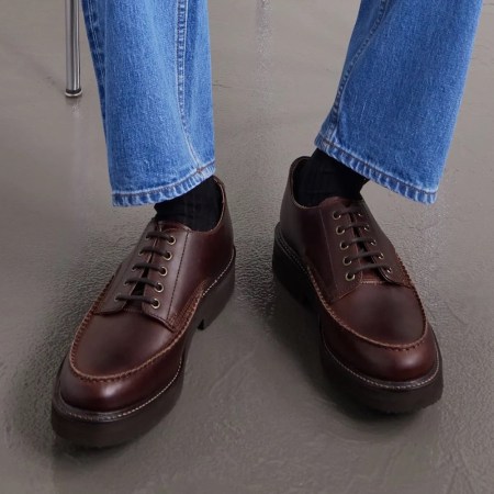A person's legs in blue jeans and black socks, wearing brown leather lace-up shoes with thick soles.