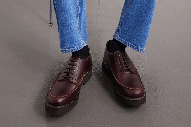 A person's legs in blue jeans and black socks, wearing brown leather lace-up shoes with thick soles.