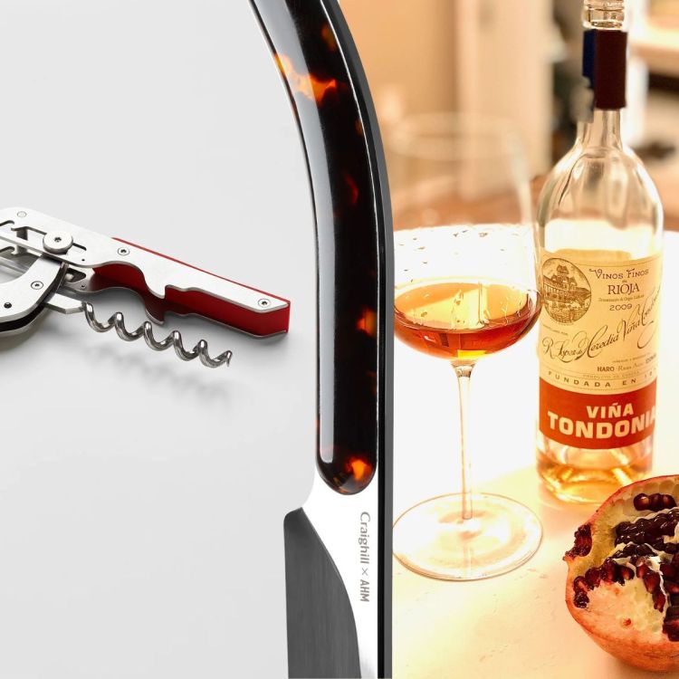 A wine key with a red handle on the left, a Champagne saber in the middle, and on the right a table with a bottle of wine, a wine glass and a pomegranate cut in half to show the seeds