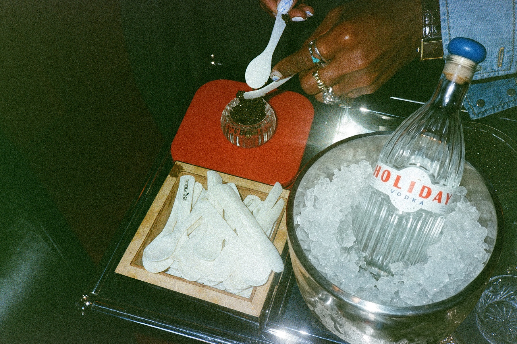 a man's hand scooping caviar into a small bowl next to a silver ice bucket with a bottle of holiday vodka