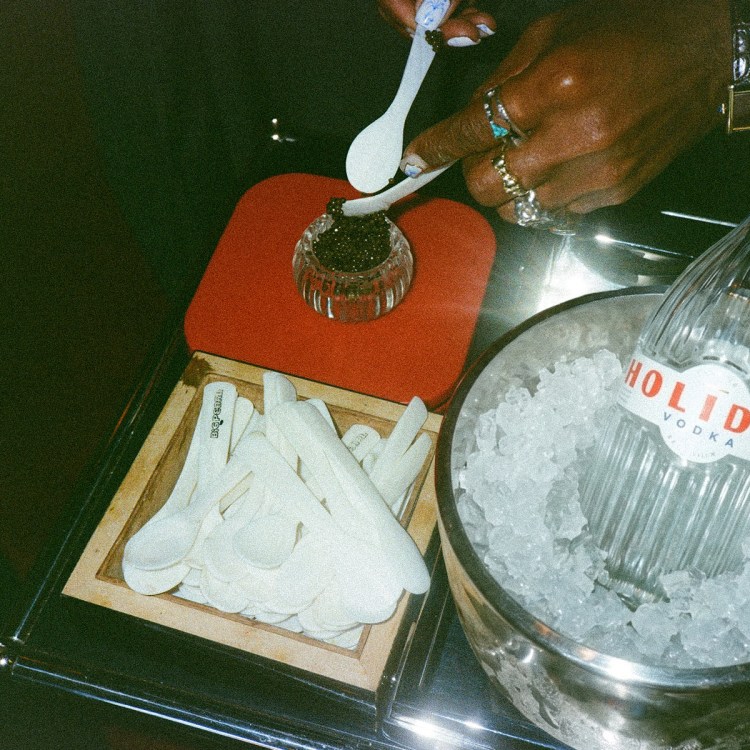 a man's hand scooping caviar into a small bowl next to a silver ice bucket with a bottle of holiday vodka