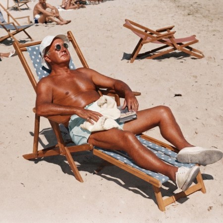 An old man relaxes shirtless at the beach with sunglasses and a beach shoes on while laying on a beach chair.