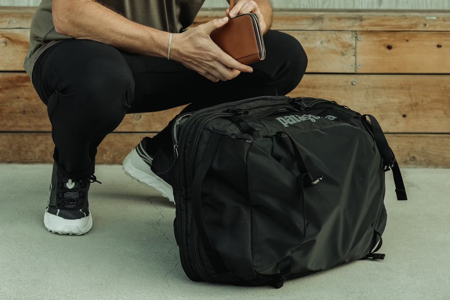 a model holding a leather wallet squatting by a black Patagonia bag.