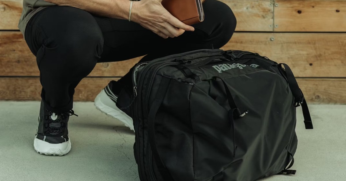 a model holding a leather wallet squatting by a black Patagonia bag.