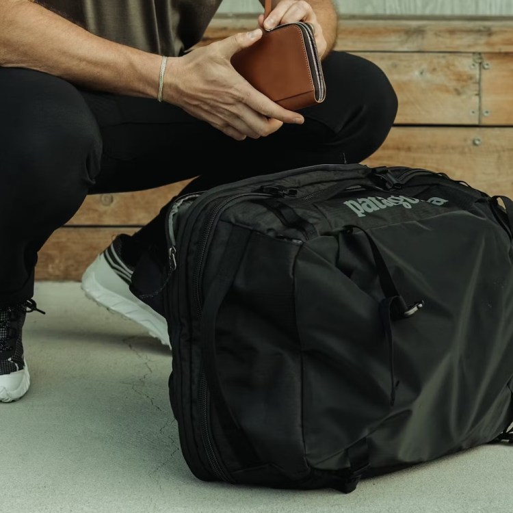 a model holding a leather wallet squatting by a black Patagonia bag.