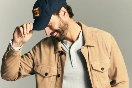A model wearing a tan suede jacket, grey henley shirt and baseball cap