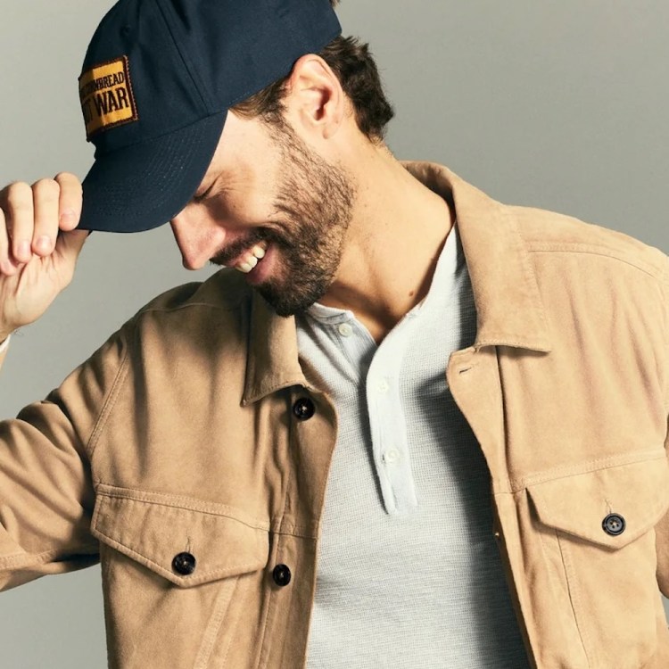 A model wearing a tan suede jacket, grey henley shirt and baseball cap