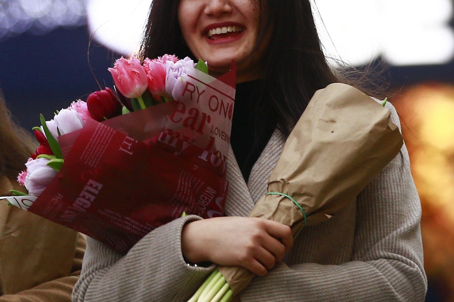 A young woman in a beige coat smiles, holding two bouquets: one of pink and red tulips, the other wrapped in brown paper.