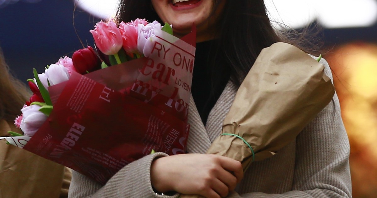 A young woman in a beige coat smiles, holding two bouquets: one of pink and red tulips, the other wrapped in brown paper.