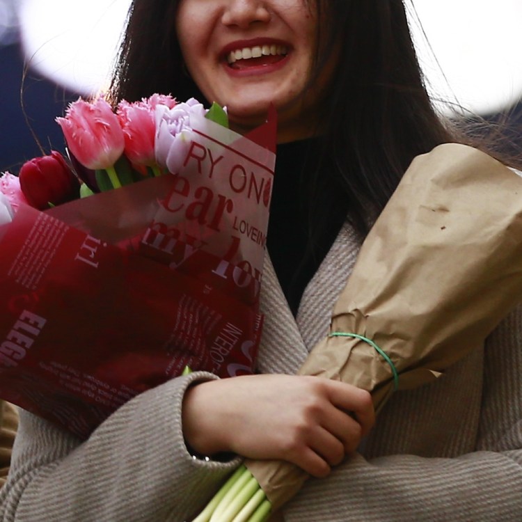 A young woman in a beige coat smiles, holding two bouquets: one of pink and red tulips, the other wrapped in brown paper.
