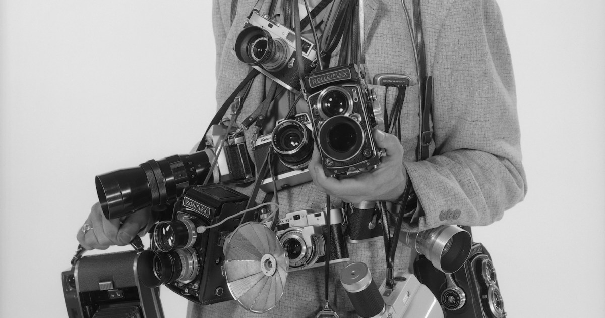 a man holding many old cameras against a white background.