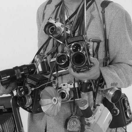 a man holding many old cameras against a white background.