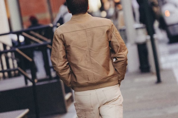A person in a tan jacket and light pants walks away from the viewer on a city street.