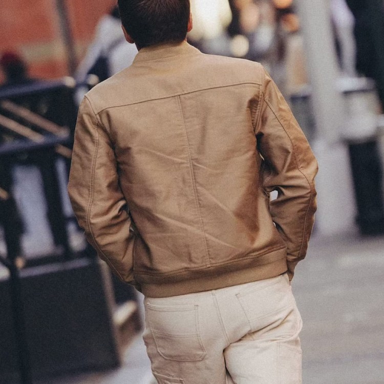 A person in a tan jacket and light pants walks away from the viewer on a city street.
