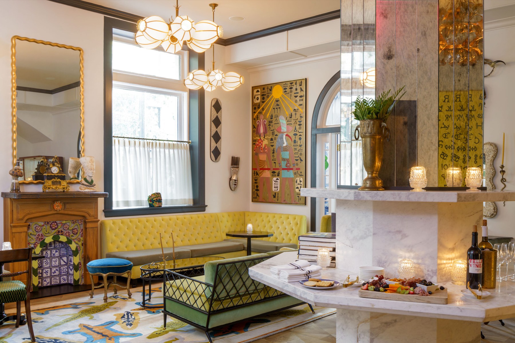 a colorful hotel lobby with yellow and green couches, a decorative fireplace and wine and charcuterie on a mirrored marble column