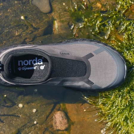 A gray and black Norda shoe with "Dyneema" branding rests in shallow water next to rocks and green algae.
