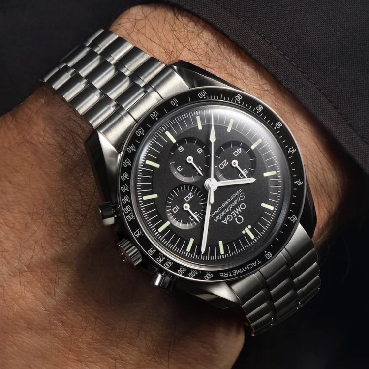 A man's wrist wearing an Omega Speedmaster Professional watch with a black dial and steel bracelet.