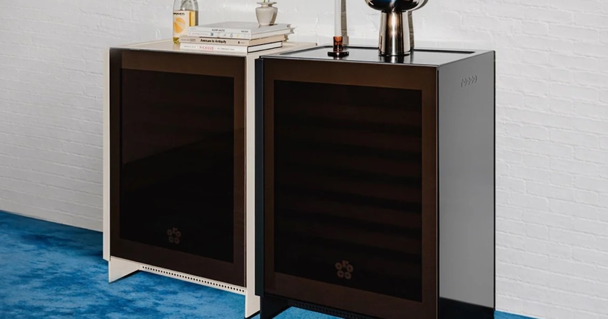 Two modern wine refrigerators, one white and one black, stand side-by-side on a blue carpet against a white brick wall.