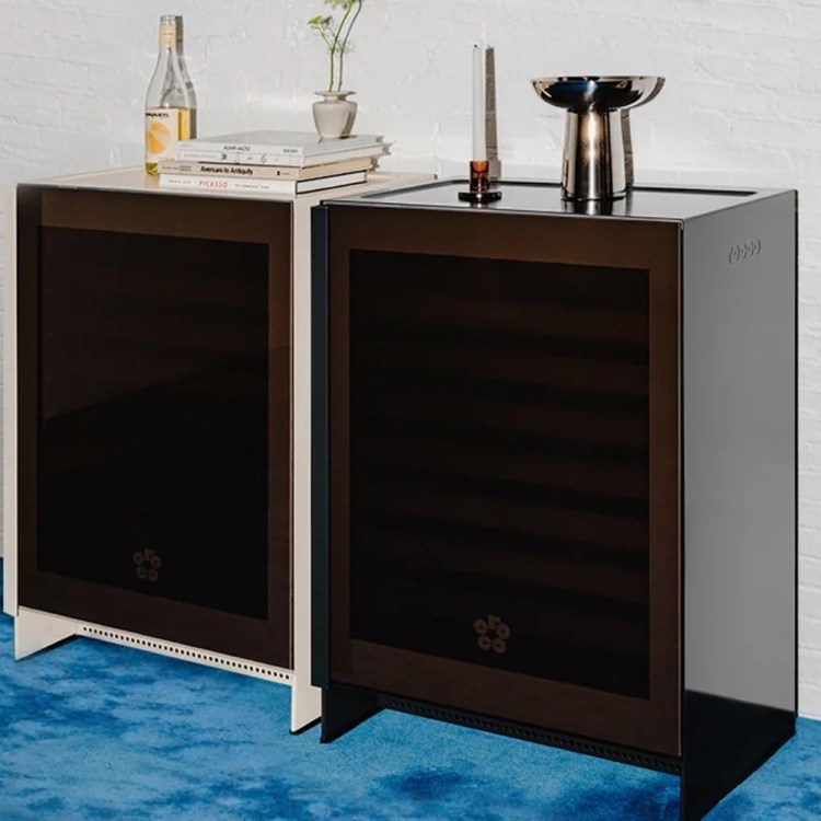 Two modern wine refrigerators, one white and one black, stand side-by-side on a blue carpet against a white brick wall.