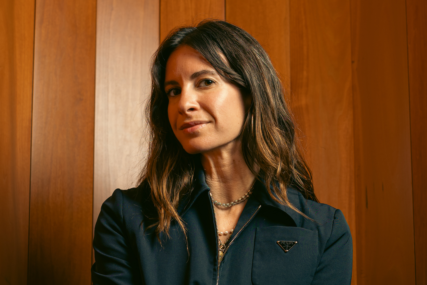 ariel arce sitting in front of a wood-paneled wall wearing a dark blue prada jumpsuit