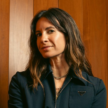 ariel arce sitting in front of a wood-paneled wall wearing a dark blue prada jumpsuit
