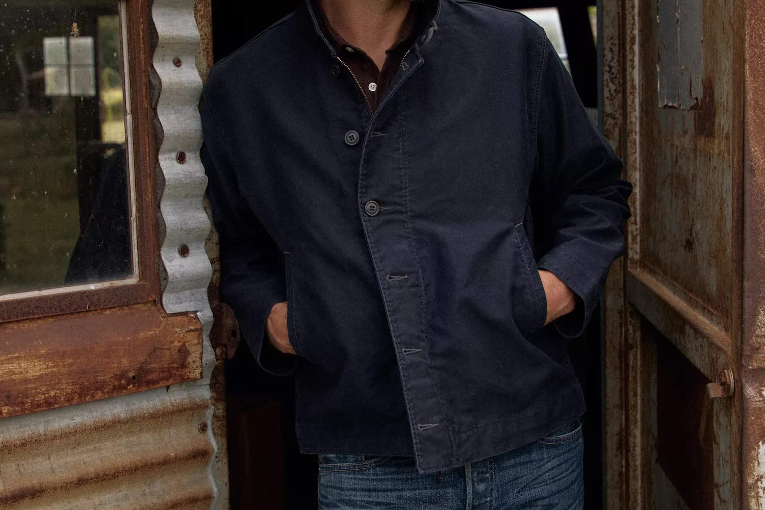 Man in a dark blue jacket and jeans with hands in pockets, leaning against a rustic corrugated metal shed.