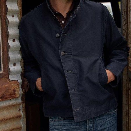 Man in a dark blue jacket and jeans with hands in pockets, leaning against a rustic corrugated metal shed.