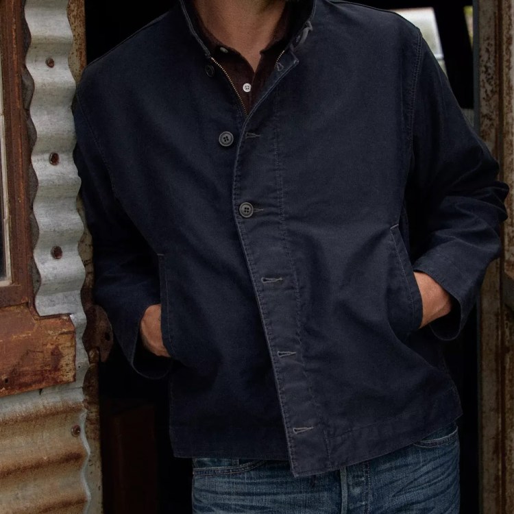 Man in a dark blue jacket and jeans with hands in pockets, leaning against a rustic corrugated metal shed.
