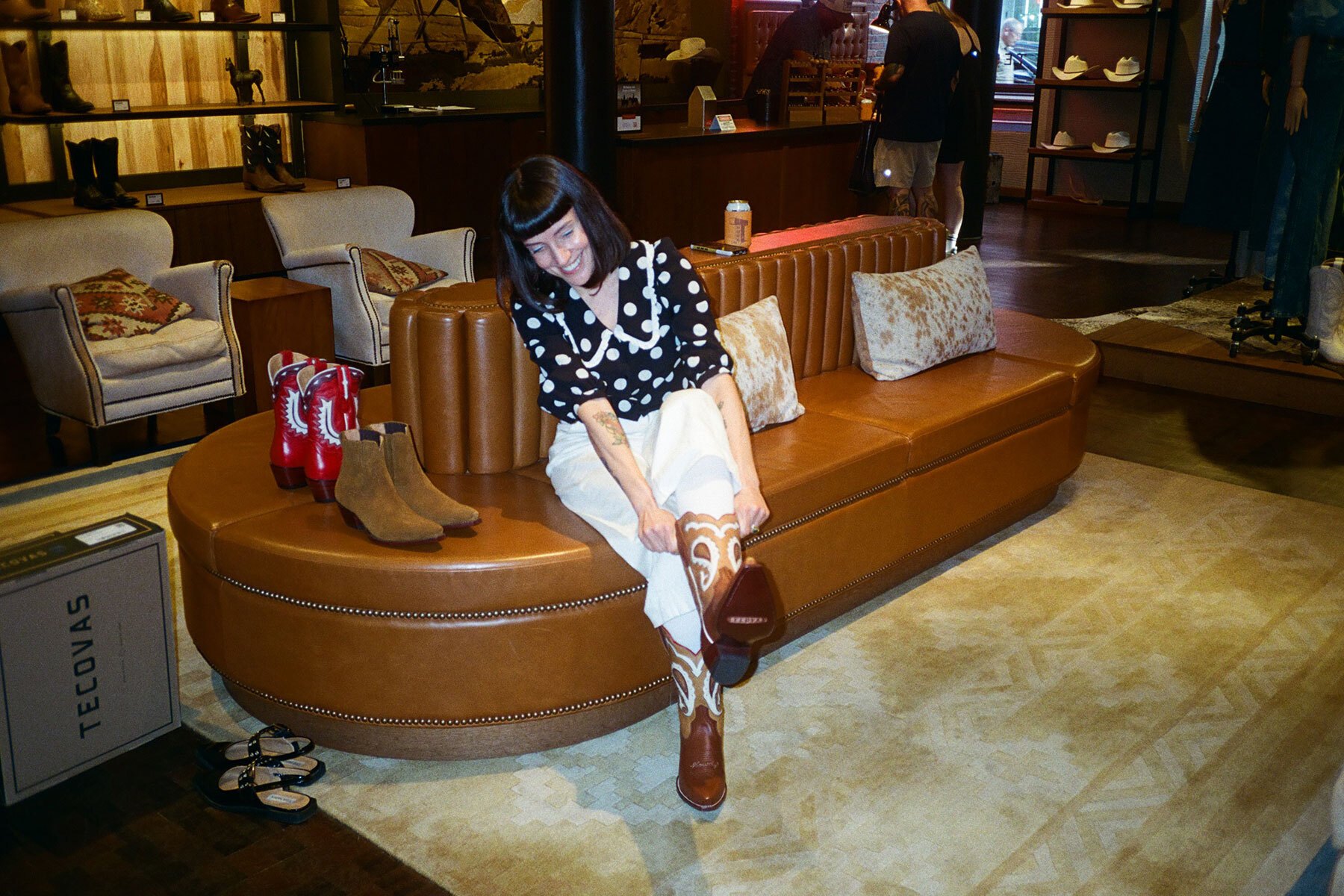a woman in a black and white polka dot shirt and white pants sitting on a brown leather couch trying on cowboy boots