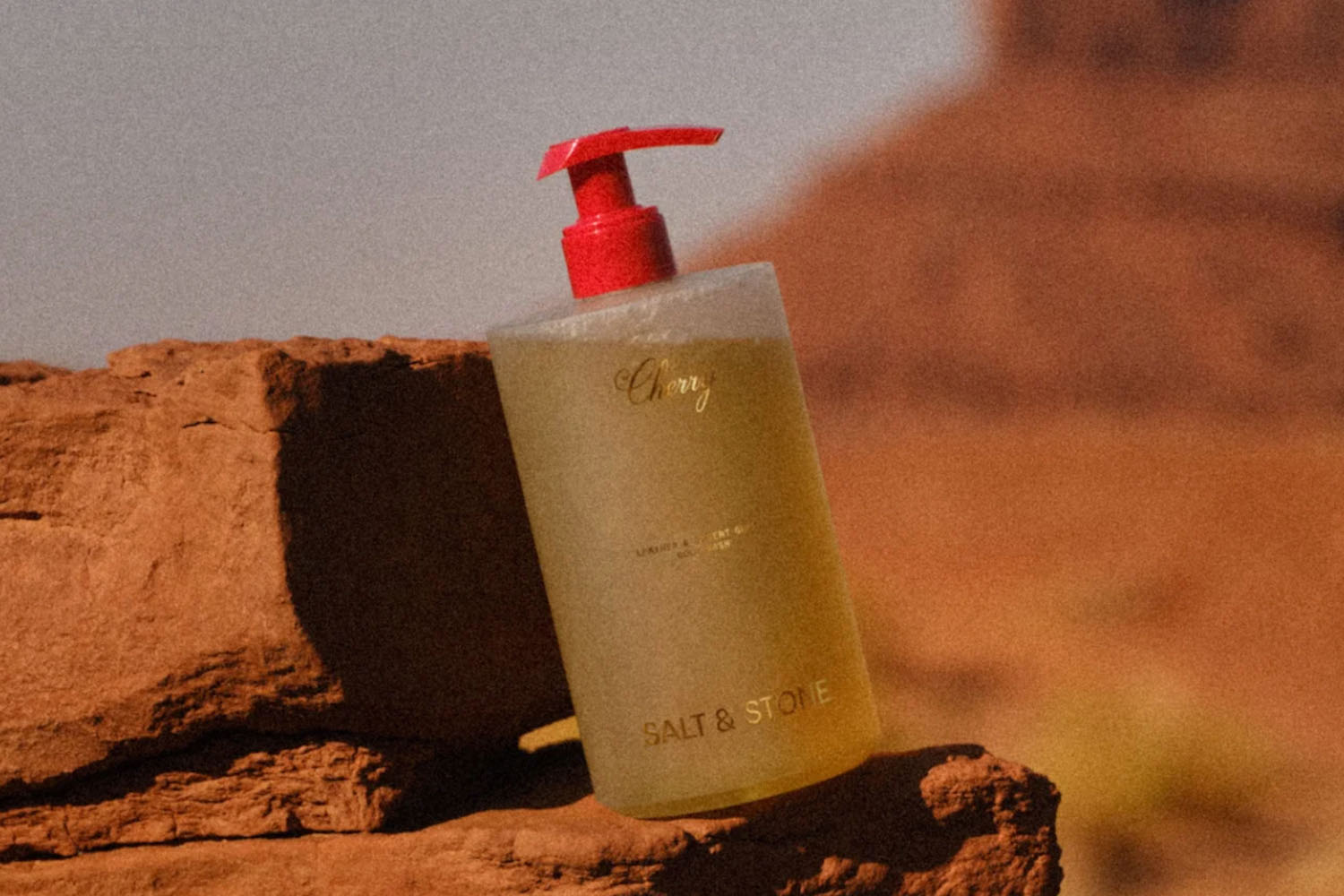 a bottle of body wash with a red cap on a rock in the desert.