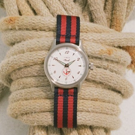 Silver watch with a cream face, red anchor logo, and a red and navy striped fabric strap.